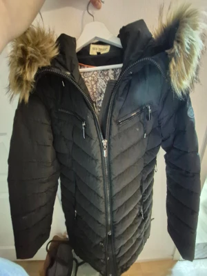 Svart lång dunjacka med fuskpäls - Snygg svart dunjacka från Coco Bonacci med långa ärmar, dragkedjor framtill och på bröstet samt huva med fuskpäls. Jackan har ett quiltat mönster och dragsko i midjan för en schysst passform. Perfekt för kalla vinterdagar.