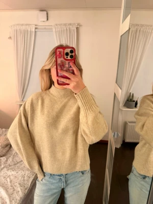 Beige stickad tröja med hög krage - Mysig beige stickad tröja med hög krage och breda ribbade muddar vid ärmslut och nederkant. Tröjan har en avslappnad passform och är perfekt att styla med jeans för en enkel och trendig look.