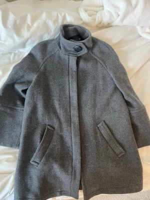 Zara Grå ullkappa - Superfin grå kappa i 64% ull med hög krage och dold knäppning. Kappan har två snedställda fickor framtill och vida trekvartsärmar. Perfekt för kyliga dagar och ger en clean look.