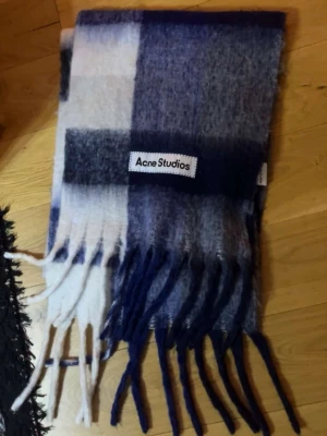  Acne Studios  - Säljer en fluffig och mjuk halsduk från Acne Studios i rutigt mönster med blå, vit och grå ton. Halsduken har långa fransar i ändarna och en tydlig Acne Studios-logga. Perfekt accessoar för att lyfta din outfit.