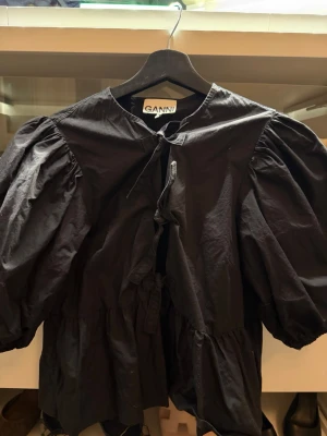 Svart voluminös blus från Ganni - Trendig svart blus från Ganni med voluminösa puffärmar och knytning framtill. Blusen har en lös passform och är tillverkad i ett lätt tyg som ger en cool och avslappnad look. Perfekt för dig som gillar statement-plagg och vill sticka ut.