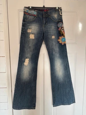 Utsvängda jeans med brodyr Desigual - Unika blå jeans med utsvängda ben, slitna detaljer och färgglad brodyr med blommor och mönster på ena benet och bakfickan. Jeansen har låg midja och dekorativt band upptill. Perfekta för dig som vill sticka ut med din stil.              Midjemått - 40 cm.  innerbenslängden - 84 cm. Jeansen är inte så strechiga, size 26