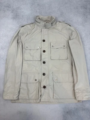 Beige vindjacka från Moncler - Stilren beige vindjacka från Moncler med fyra framfickor med lock och knappar, hög krage och knappslå. Jackan har en diskret logotyp på insidan och är tillverkad i ett lätt, vindtätt material. Perfekt för dig som vill ha en klassisk och funktionell look.