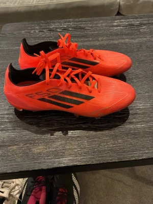Röda Adidas fotbollsskor med svarta detaljer - Säljer ett par snygga röda Adidas fotbollsskor med svarta ränder på sidan och silvriga Adidas-loggor bak. Skorna har snörning och låg profil, perfekt för snabba rörelser på planen. Ovandelen är i syntetmaterial och insidan är svart. Köpta för 2000kr och använda max 5-6 ggr då jag växte ur dom
