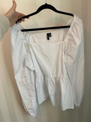 Vit blus med pärldetaljer - Söt vit blus från VERO MODA med långa ärmar och dekorativa pärlrosetter vid axlarna. Blusen har en luftig passform och rynkade detaljer vid midjan. Använd 2-3 gånger.