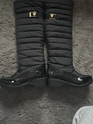 Svarta knähöga stövlar med gulddetalj - Snygga svarta knähöga stövlar från Versace med quiltad textil på skaftet och lackad finish på foten. Dekorerade med guldfärgade spännen med Versace-logga upptill. Perfekta för att lyfta din outfit med en lyxig touch. Bought in schweitz🇨🇭 