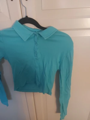 Turkos ribbad långärmad polotopp - Snygg turkos ribbad polotopp med lång ärm och klassisk krage. Toppen har knappar framtill och är croppad i modellen, perfekt för en trendig look. Materialet känns mjukt och stretchigt.