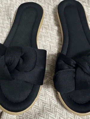 Svarta sandaler med knutdetalj - Säljer ett par svarta sandaler med breda tygband och snygg knutdetalj framtill. Sulan är beige och har en lätt platå. Perfekta för sommaren och riktigt bekväma att glida i. Materialet på ovandelen är mjukt tyg och sulan är i syntet.