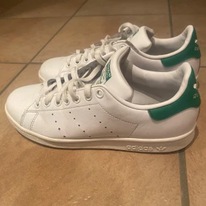Adidas Stan Smith vita sneakers - Klassiska vita Adidas Stan Smith sneakers med gröna detaljer på häl och plös. Skorna har lågt skaft, perforerade ränder på sidorna och är tillverkade i slätt läder. Ikonisk logga på hälen och tungan. Perfekta för en clean och tidlös stil.