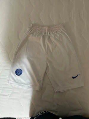 Vita PSG shorts från Nike - Säljer ett par vita PSG shorts från Nike med blå logga och klubbmärke. Shortsen har elastisk midja och är tillverkade i ett lätt och bekvämt polyestermaterial, perfekta för träning eller fotboll.