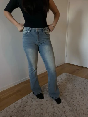 Bootcut ljusblå jeans med hög midja - Säljer ett par ljusblå bootcut jeans med hög midja och klassisk femficksdesign. Jeansen har en snygg tvätt med slitningar framtill och är tillverkade i ett mjukt denimtyg som sitter bekvämt. Perfekta för en avslappnad och trendig look.