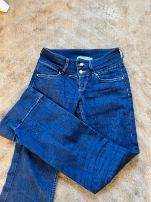 Mörkblå bootcut jeans med låg midja - Säljer ett par mörkblå bootcut jeans med låg midja och två knappar i gylfen. Klassisk femficksmodell i stretchigt denimtyg som sitter snyggt och bekvämt. Perfekta för dig som gillar en retroinspirerad look med utsvängda ben.