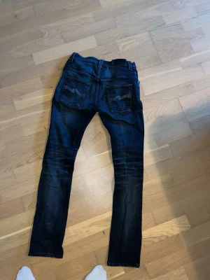 Mörkblå skinny jeans från Nudie - Säljer ett par mörkblå skinny jeans från Nudie med klassiska fem fickor och snygga slitningar på benen. Jeansen har kontrastsömmar och Nudie-loggan broderad på bakfickorna. Tillverkade i stretchigt denim för skön passform.