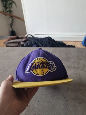 Lakers snapback keps - Snygg snapback keps i lila och gult med Los Angeles Lakers broderad logga framtill och Mitchell & Ness broderat bak. Klassisk platt skärm och justerbar storlek. 