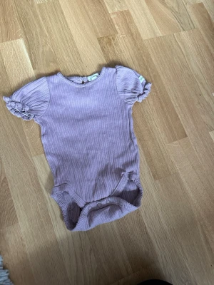 Ljuslila ribbad body med puffärm - Supersöt ljuslila body i ribbad bomull med korta puffärmar och volangkant. Rund halsringning med liten knapp i nacken och tryckknappar i grenen för enkel påklädning. Perfekt för en gullig och bekväm look.