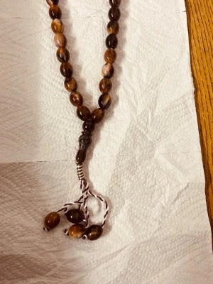 Brun bönkedja med pärlor - Säljer en snygg bönkedja med ovala pärlor i olika bruna nyanser och fina ådringar. Pärlorna är trädda på en vit och brun tvinnad tråd och avslutas med dekorativa hängande pärlor. Perfekt accessoar för dig som gillar unika detaljer.