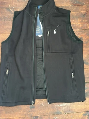 Svart väst från Polo Ralph Lauren - Svart väst med dragkedja från Polo Ralph Lauren. Västen har ståkrage, tre fickor med dragkedja och meshfoder på insidan. Klassisk logga broderad på bröstet. Perfekt för lager-på-lager och stilren look.