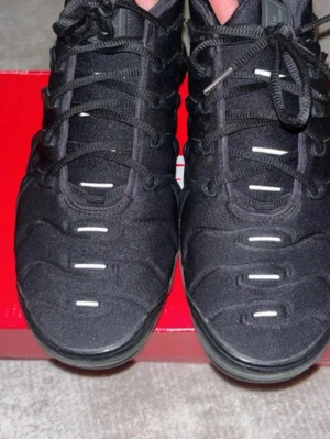 Svarta Nike Air Vapormax Plus sneakers - Snygga svarta Nike Air Vapormax Plus sneakers med unik design och synliga luftbubblor i sulan. Skorna har svarta snören, diskreta vita detaljer och en sportig look. Ovandelen är i textil och syntet, perfekt för dig som gillar streetwear.En balång är paff men går att fixa ! 