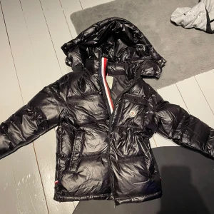 Moncler jacka - Snygg svart pufferjacka från Moncler med huva och dragkedja. Jackan har ett quiltat mönster och en färgglad detalj vid dragkedjan. Perfekt för kyliga dagar. Är i storlek M men passar även S