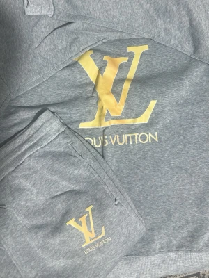 Grått Louis Vuitton set med logga - Säljer ett grått set från Louis Vuitton med stor gul LV-logga och text på både överdel och underdel. Setet är gjort i mjukt bomullsmaterial och har en avslappnad passform. Perfekt för dig som gillar exklusiva märken och stilren design.