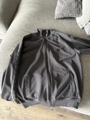 Grå track jacket från Fred Perry - Säljer en grå track jacket från Fred Perry med dragkedja framtill. Jackan har diskret logga på bröstet och mönstrade band längs ärmarna. Perfekt för en sportig och avslappnad stil. Fast pris