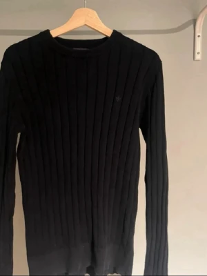 Svart ribbstickad tröja Morris - Snygg svart ribbstickad tröja från Morris med rund halsringning och diskret logga på bröstet. Tröjan har långa ärmar och är tillverkad i ett mjukt, stretchigt material som sitter tajt mot kroppen. Perfekt för dig som gillar stilrena och klassiska plagg.