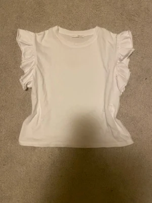 Vit topp med volangärmar från H&M - Säljer en söt vit topp från Zara med volangdetaljer på ärmarna. Toppen har rund halsringning och är tillverkad i mjuk bomull, perfekt för en fräsch och enkel look. också menad som en sommartröja och är i storlek S 💗💗