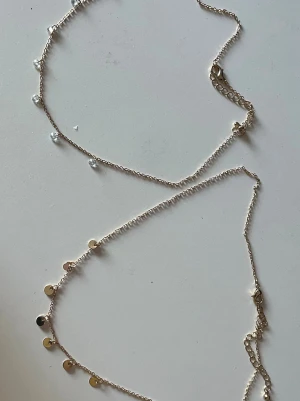 Två guldhalsband med detaljer - Säljer två halsband i guldfärgad metall. Det ena halsbandet har små vita ” kristall ” pärlor längs kedjan, medan det andra har små runda guldfärgade plattor som dekoration. Perfekta för att kombinera eller bära var för sig för en trendig look.