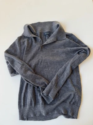 Grå stickad tröja från Riley - Snygg grå stickad tröja från Riley i mjuk merinoull. Tröjan har krage och knäppning med två knappar vid halsen. Skriv ifall du har några frågor!