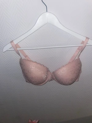 Rosa spets-bh med vadderade kupor - Söt ljusrosa bh med vadderade kupor och spetsdetaljer. Justerbara axelband och hakknäppning bak. Kuporna har en lätt push-up-effekt och kanten är dekorerad med vågig spets. Perfekt för dig som gillar romantisk och feminin stil.