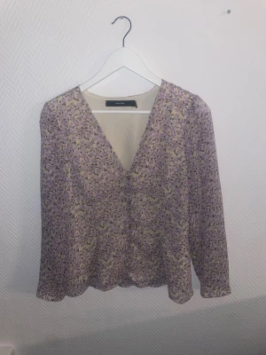 Lila blommig blus från Vero Moda - Söt blus från Vero Moda i lila och beige med småblommigt mönster. Blusen har v-ringning och långa ärmar i ett lätt, transparent material som ger en luftig känsla. Perfekt att styla med jeans eller kjol för en trendig look.