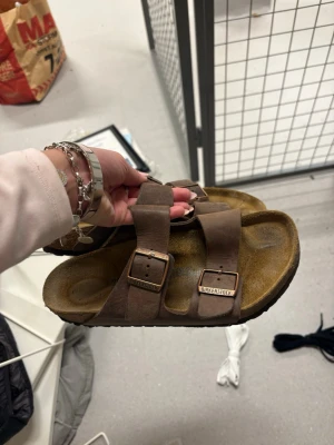 Bruna Birkenstock sandaler i läder - Säljer ett par klassiska bruna Birkenstock sandaler med två justerbara remmar och metallspännen. Tillverkade i läder med en ergonomisk fotbädd för extra komfort. Perfekta för dig som gillar en avslappnad och tidlös stil.