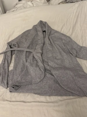 Ljusgrå omlottkofta med knytband - Mysig ljusgrå omlottkofta med långa ärmar och bred krage. Koftan har ett matchande knytband i midjan för en avslappnad och snygg look. Perfekt att slänga på sig över en t-shirt eller linne. Materialet känns mjukt och stretchigt.