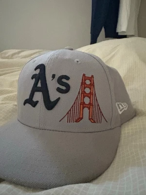 Grå New Era keps med brodyr - Snygg grå keps från New Era med broderad A's-logga och Golden Gate-bron på framsidan. På baksidan finns en liten broderad drink och MLB-logga. Klassisk rak skärm och strukturerad passform, perfekt för dig som gillar streetwear och sportig stil.
