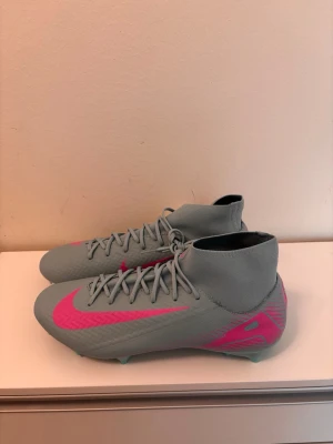 Nike grå-rosa fotbollsskor med socka - Snygga fotbollsskor från Nike i ljusgrått med rosa detaljer och klassisk swoosh. Skorna har en strumpliknande hög krage för extra stöd runt ankeln och snörning framtill. Tillverkade i syntetmaterial för bra grepp och kontroll på planen.