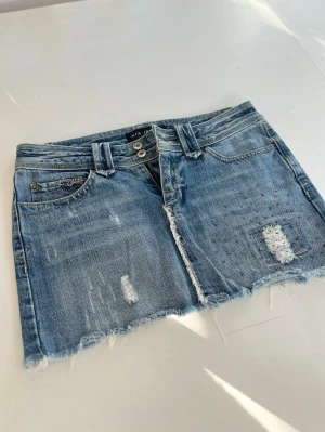 Blå jeanskjol med slitningar från Nix jeans - Säljer en blå jeanskjol från Nix jeans  med rå kant och slitna detaljer framtill och baktill. Kjolen har klassiska fem fickor, bälteshällor och knappgylf. Perfekt för dig som gillar en avslappnad och trendig stil. Mått på bilden