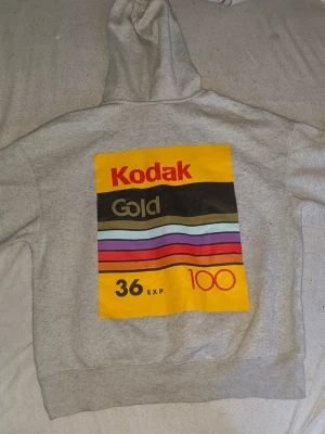 Grå hoodie med Kodak Gold-tryck - Snygg grå hoodie med stor färgglad Kodak Gold-etikett på ryggen och mindre tryck framtill. Hoodien har huva och känguruficka. Materialet är mjukt och bekvämt, perfekt för dig som gillar retrostil och streetwear. Flera färger i trycket: gul, röd, svart, lila och blå.