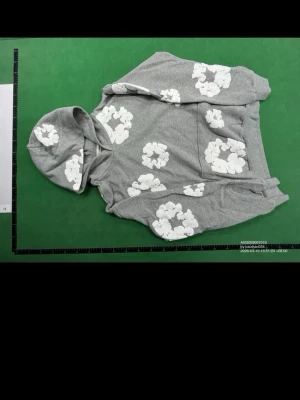 Grå hoodie med vita blommor Denim Tears - Snygg grå hoodie från Denim Tears med stora vita blommor tryckta över hela plagget. Hoodien har en klassisk huva och känguruficka framtill. Tillverkad i mjukt bomullsmaterial som känns skönt mot huden. Perfekt för dig som gillar streetwear och unika prints.