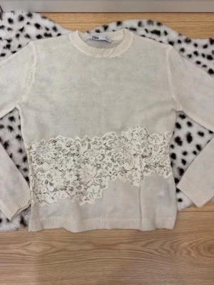 Vit stickad tröja med spets från Zara - Supersöt vit stickad tröja från Zara med bred spetsdetalj över magen. Tröjan har rund halsringning, långa ärmar och ribbade muddar. Materialet är mjukt och lätt, perfekt för lager på lager. Spetsen ger en romantisk och trendig touch.