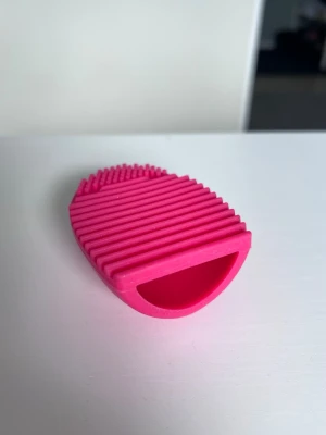 Rosa borstrengörare i silikon, oanvänd - Praktisk borstrengörare i rosa, tillverkad av mjukt silikon. Räfflad och knottrig yta för effektiv rengöring av sminkborstar. Träs på fingrarna för vid användning.