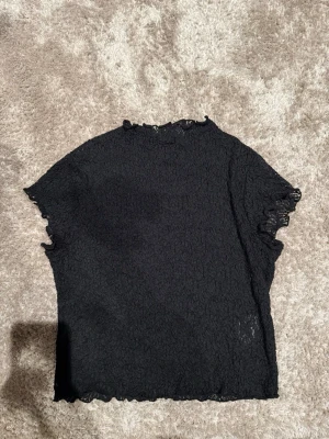 Svart spetsig topp från C&A - Svart topp från H&m med korta ärmar och vågiga kanter. Tunt spetsliknande material som ger en cool och trendig look. Perfekt att styla med jeans eller kjol för en snygg outfit.