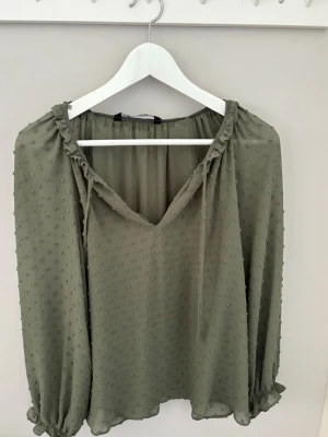 Grön Zara blus  - Snygg olivgrön blus med v-ringning och volangkant vid halsen. Blusen har ett hål vid utingincen men man märker inte alls av det.  Tillverkad i ett tunt, transparent tyg som ger en avslappnad och trendig look.