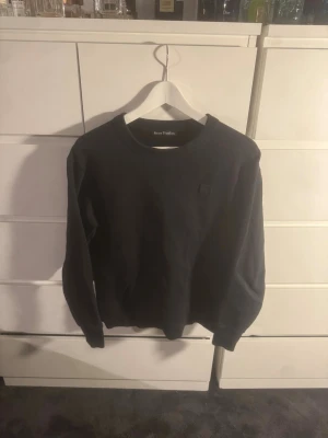 Svart sweatshirt från Acne Studios - Svart sweatshirt från Acne Studios med rund hals och diskret logopatch på bröstet. Tröjan har långärmade ärmar och ribbade muddar vid hals, ärmslut och nederkant. Materialet är mjuk bomull som ger en skön och avslappnad känsla.