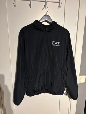 Svart vindjacka från EA7 Emporio Armani - Snygg svart vindjacka från EA7 Emporio Armani med huva och dragkedja framtill. Jackan har två sidofickor med dragkedja och logga på bröstet. Tillverkad i lätt och smidigt material som passar perfekt för blåsiga dagar. Superlitrt hål lägst ut på armen men inget man ser eller tänker på, se på bild 3