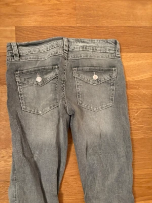 Grå bootcut jeans med ficklock - Snygga grå jeans med bootcut passform och slitna detaljer. Jeansen har två bakfickor med ficklock och knapp, samt klassiska framfickor. Materialet är denim i bomull och de har en låg midja. Perfekta för en avslappnad och trendig look.
