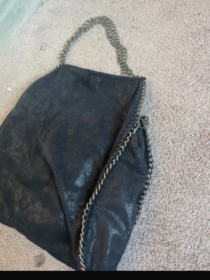 Svart handväska med kedjedetaljer - Snygg svart handväska i mjukt mockaliknande material med grova silverfärgade kedjor längs kanterna och som axelrem. Väskan har en slouchy form och ger en cool, trendig vibe. Perfekt för dig som gillar statement-accessoarer och vill sticka ut.