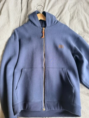 Blå zip hoodie från Miu Miu - Snygg blå hoodie från Miu Miu med dragkedja i guld och läderdetalj på zippen. Hoodien har huva, två stora fickor fram och diskret Miu Miu-logga broderad på bröstet. Tillverkad i mjukt bomullsmaterial som känns riktigt skönt mot huden.