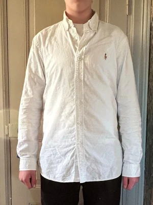 Vit skjorta från Ralph Lauren - Klassisk vit skjorta från Ralph Lauren med button-down krage och broderad logga på bröstet. Skjortan har lång ärm, knappar framtill och är tillverkad i bomull. Perfekt till en clean och stilren look.
