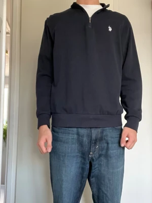 Marinblå quarter zip från U.S Polo ASSN. - Snygg marinblå långärmad tröja från U.S Polo med quarter zip och klassisk broderad logga på bröstet. Tröjan har ribbade muddar och krage, perfekt för en stilren och avslappnad look. Tillverkad i mjuk bomullsblandning.