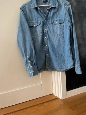 Slitet blå jeansskjorta från Gant - jeansskjorta från Gant med två bröstfickor och vita knappar. Skjortan har lång ärm och en avslappnad passform, perfekt till jeans eller chinos. Tillverkad i mjuk bomull för en bekväm känsla. Storlek 170 junior, som en small herr